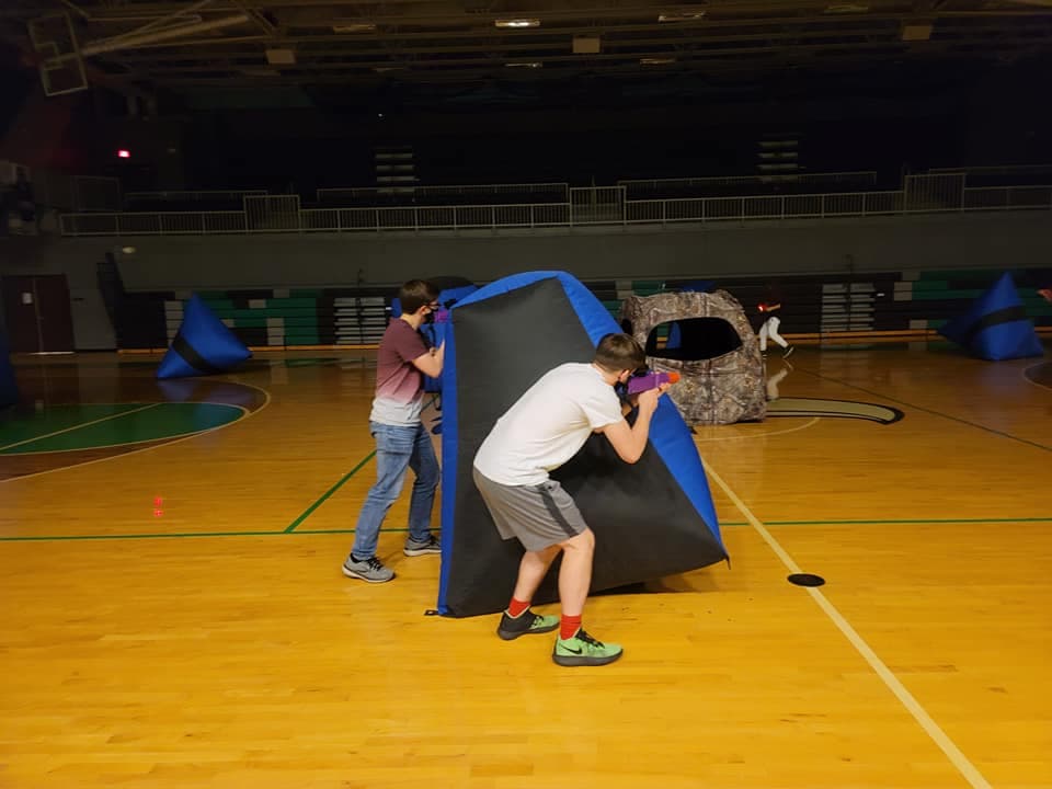 Two players taking aim with laser blasters in a game of laser tag standing near a obstacle with a camo tent in the background inside a gym