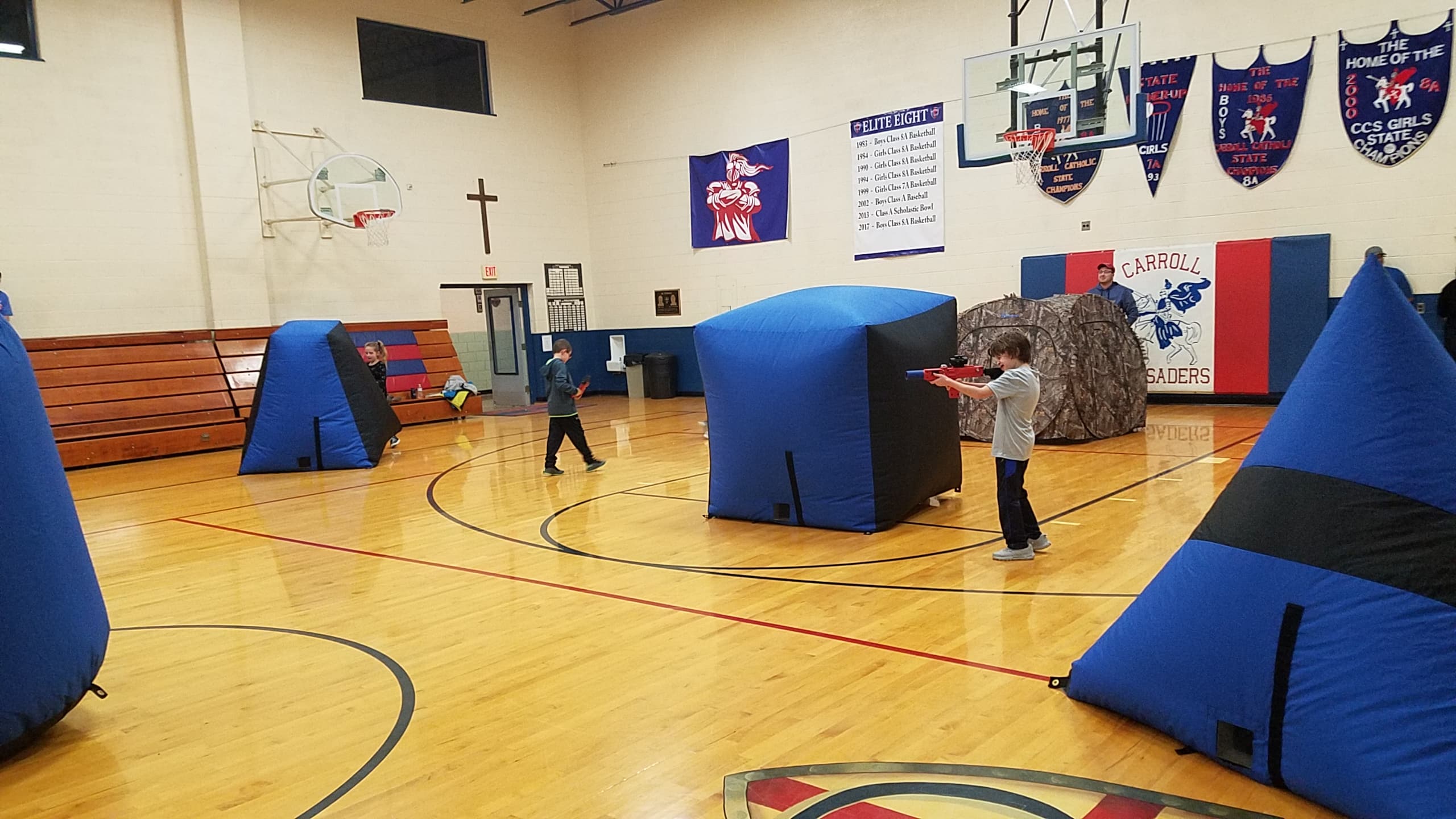 Two kids playing laser tag around black and blue obstacles inside a gym