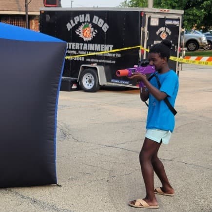 A child smile while holding laser tag blasters in a gymnasium. A basketball hoop and inflatable obstacles are visible in the background.