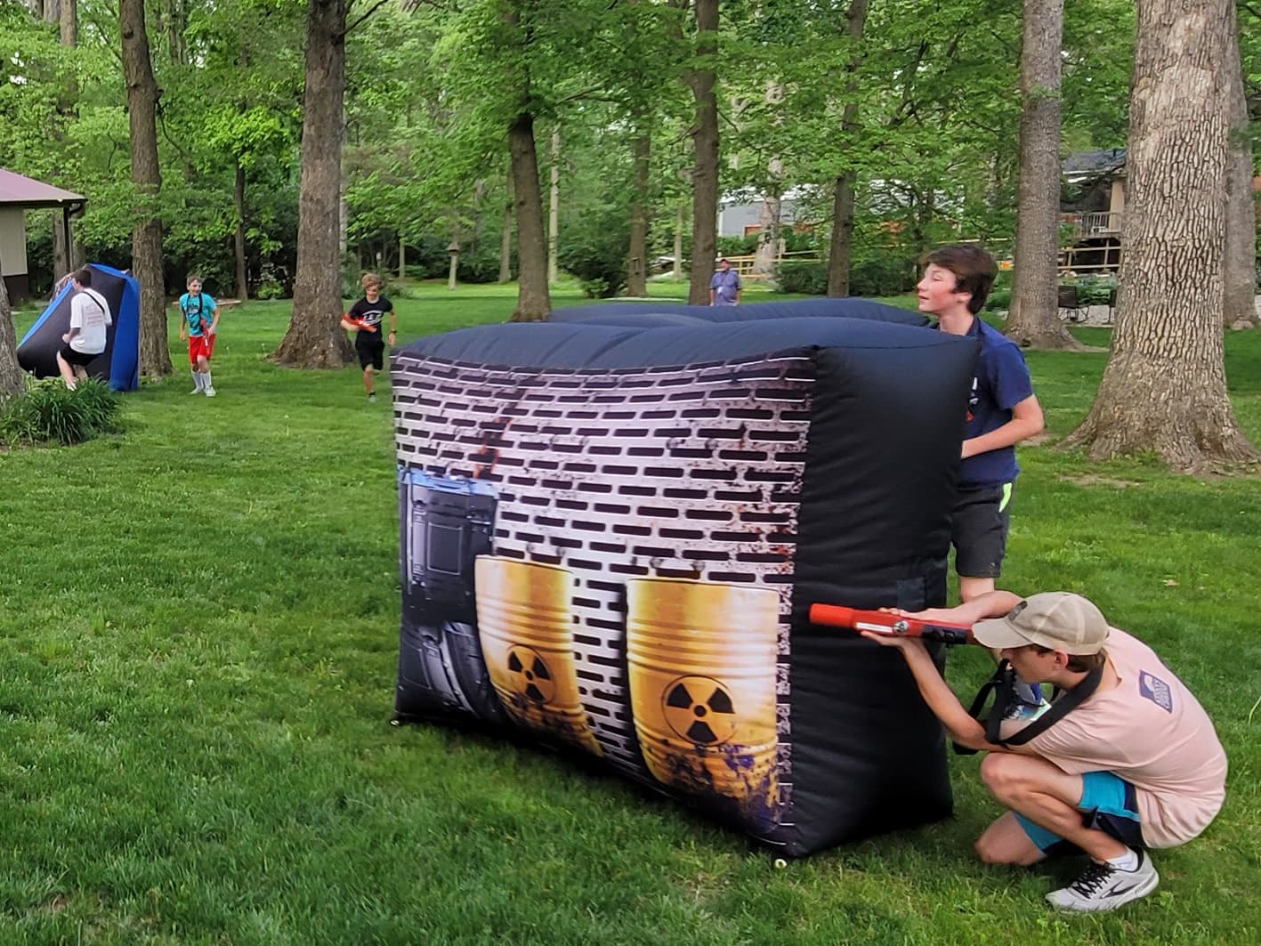 Kids play laser tag in a wooded yard, hiding behind inflatable barriers. The scene is lively and fun, with players focused and energetic.