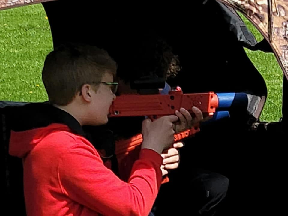 A person in a red hoodie excitedly aims a colorful laser tag blaster from inside a shaded tent, set on a vibrant green lawn.