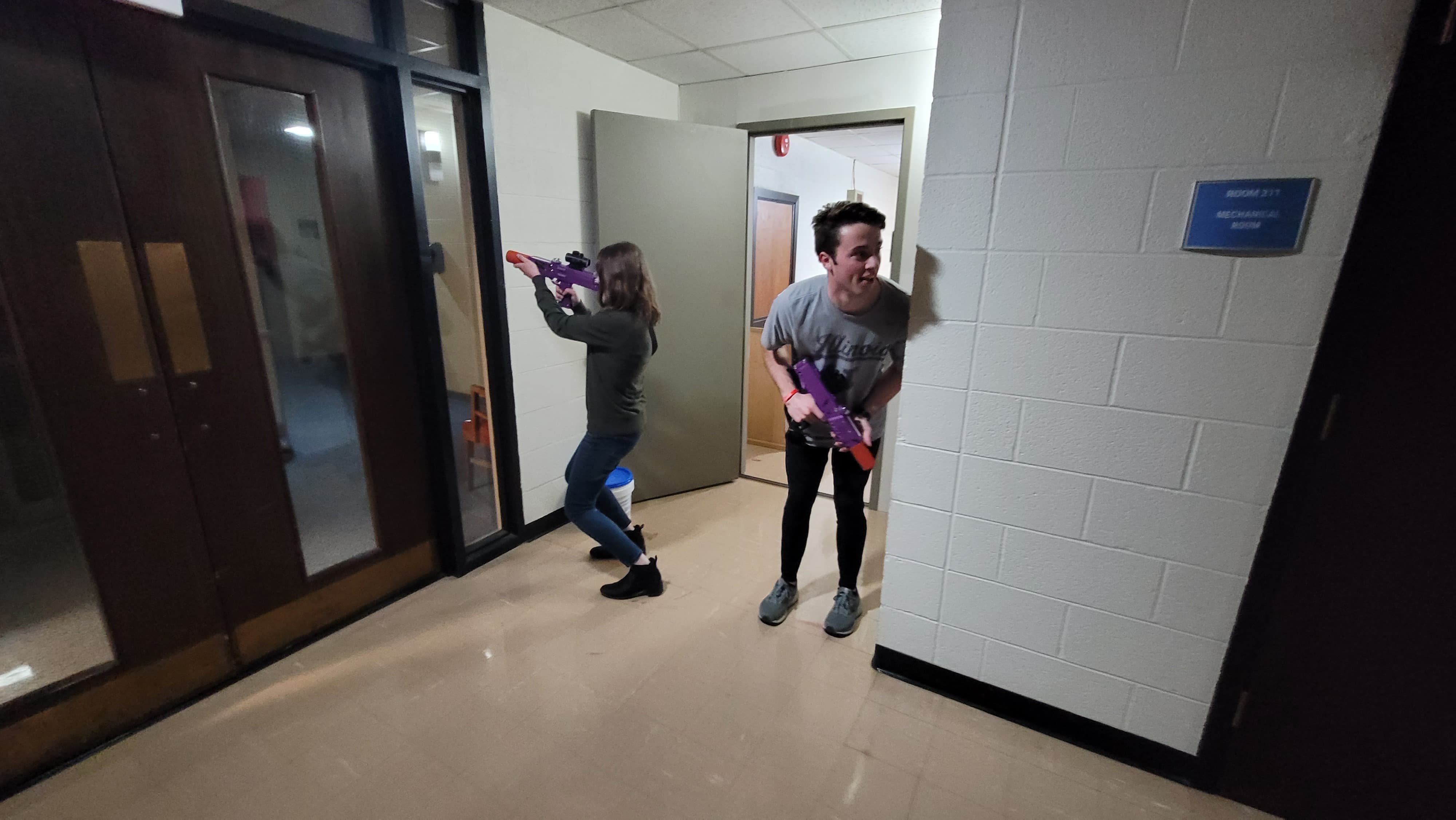 Two people in a hallway with laser tag blasters, poised for a playful ambush. One crouches around a corner; the other aims forward. The scene is lighthearted.