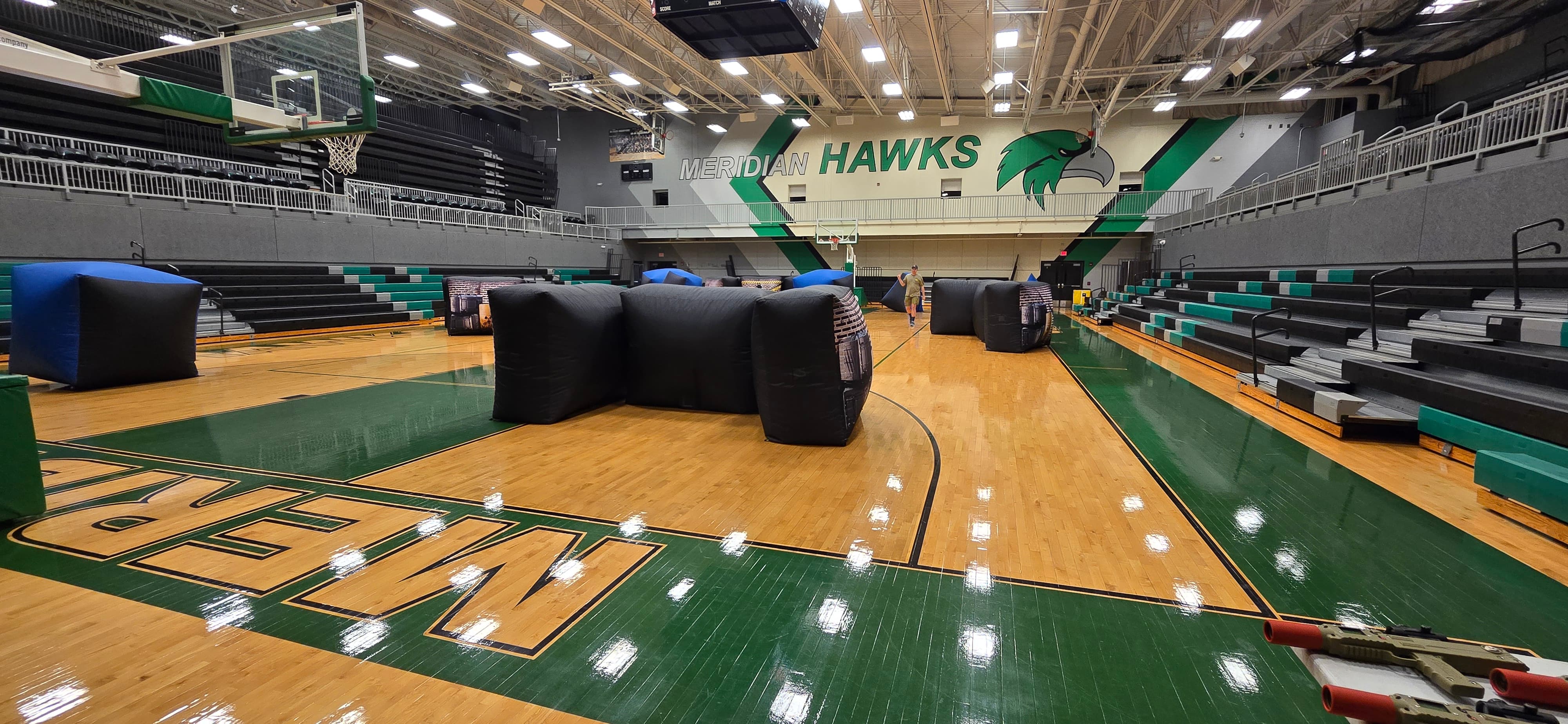 Spacious gym with inflatable barriers and laser tag blasters set up for a game. 'Meridian Hawks' is displayed on the wall with green bleachers alongside.