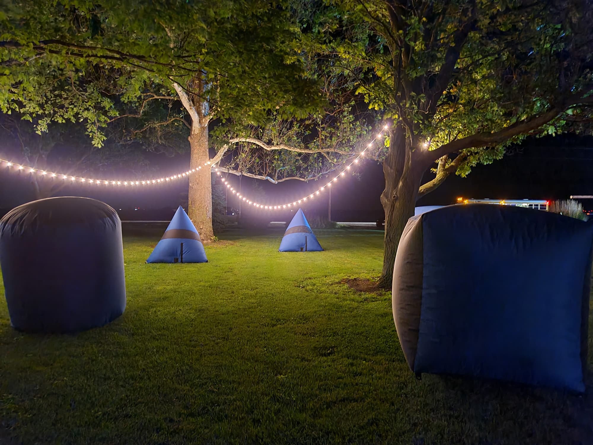 Nighttime outdoor laser tag arena with inflatable bunkers set on grass under string lights between trees.