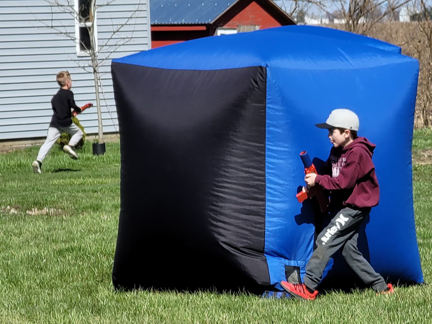 Two people in a hallway with laser tag blasters, poised for a playful ambush. One crouches around a corner; the other aims forward. The scene is lighthearted.